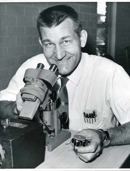 William McLellan used a microscope to inspect his tiny motor, while holding a model one million times larger. 1960 (William McLellan Special Collection)