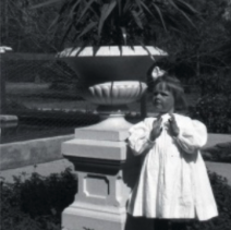 a black and white photo of a small child in a white dress next to a fountain