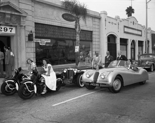 A historical image of two teenage girls on motorcycles in front of two sports cars parked in front of a Jaguar dealership in Pasadena in 1950.